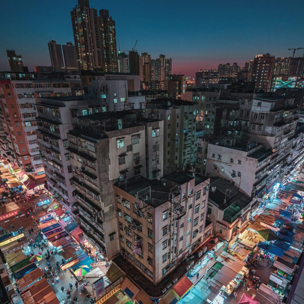 The Night Fall features a vibrant night market scene like an art print, with colorful street stalls lit brightly amidst tall residential buildings under a dark sky, set against a city skyline at dusk.