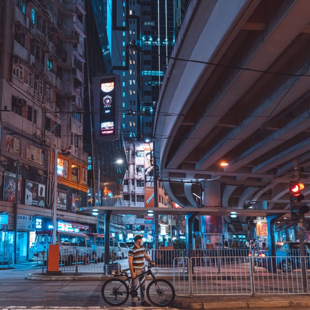 A cyclist stands with a bike at a city intersection at night, resembling an art print. The scene features a large overpass and tall buildings with bright neon signs. The cityscape glows in blue and orange tones, creating a vibrant urban atmosphere. Product Name: Cyclist.