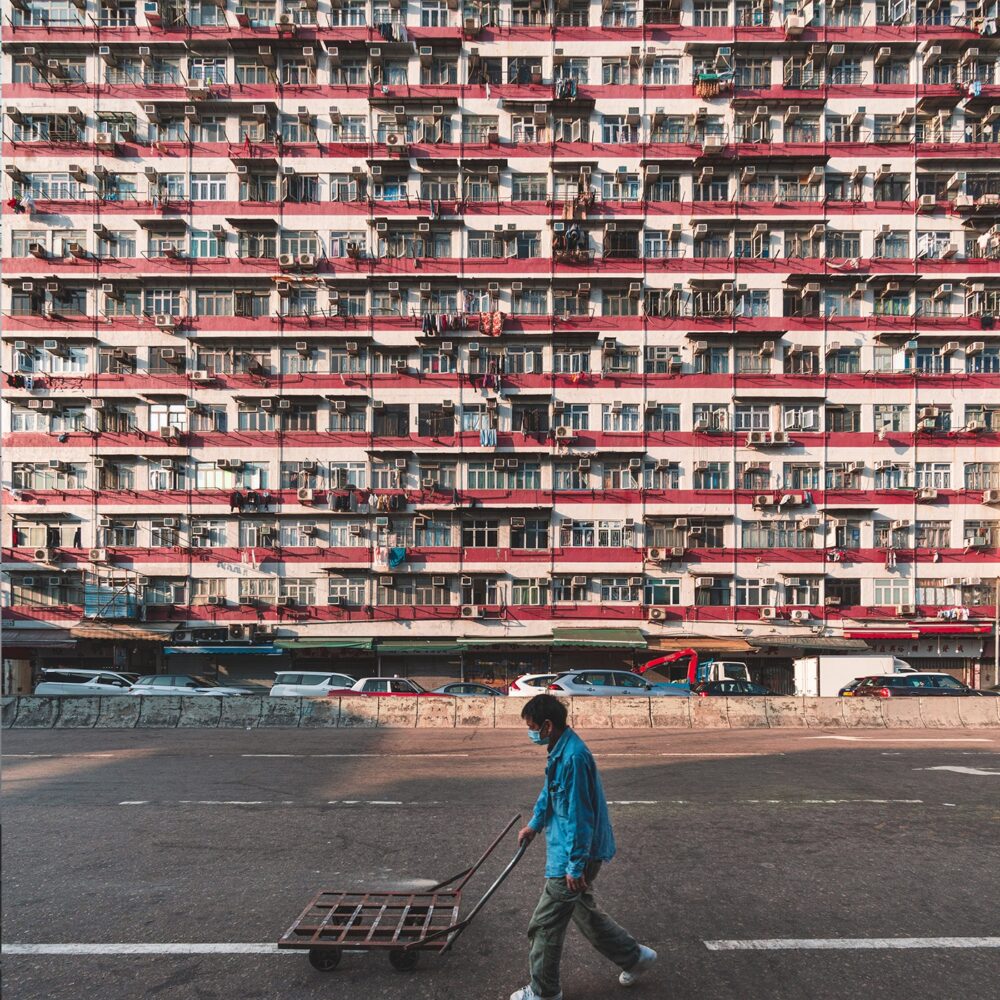Wearing a blue jacket, a person pushes a cart across an empty urban street in Petty Citizens art print. A large apartment building with balconies and AC units fills the background, while cars line the street.