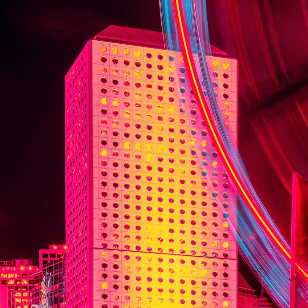 The tall buildings grid-like facade, lit in vibrant pink and yellow at night, echoes a striking art print. Blue and red light trails arc dynamically across the sky, adding motion to the urban scene—all captured in Perpetuity.