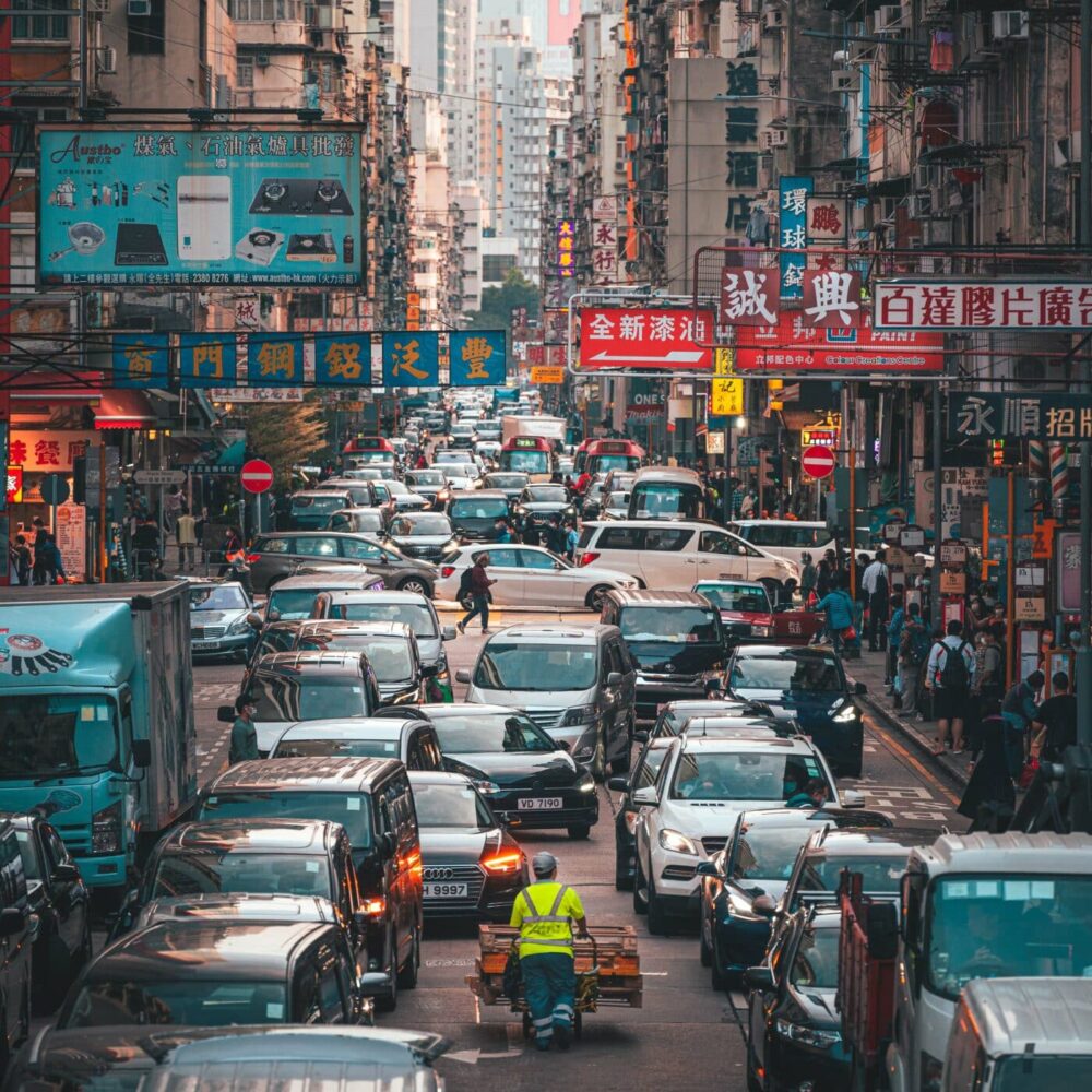 An urban street scene with heavy traffic, including cars, vans, and trucks. A person in a yellow vest rides a tricycle cart through the chaos. This vibrant depiction resembles an art print titled Traffic Jam, adorned with colorful signs and Chinese characters above.