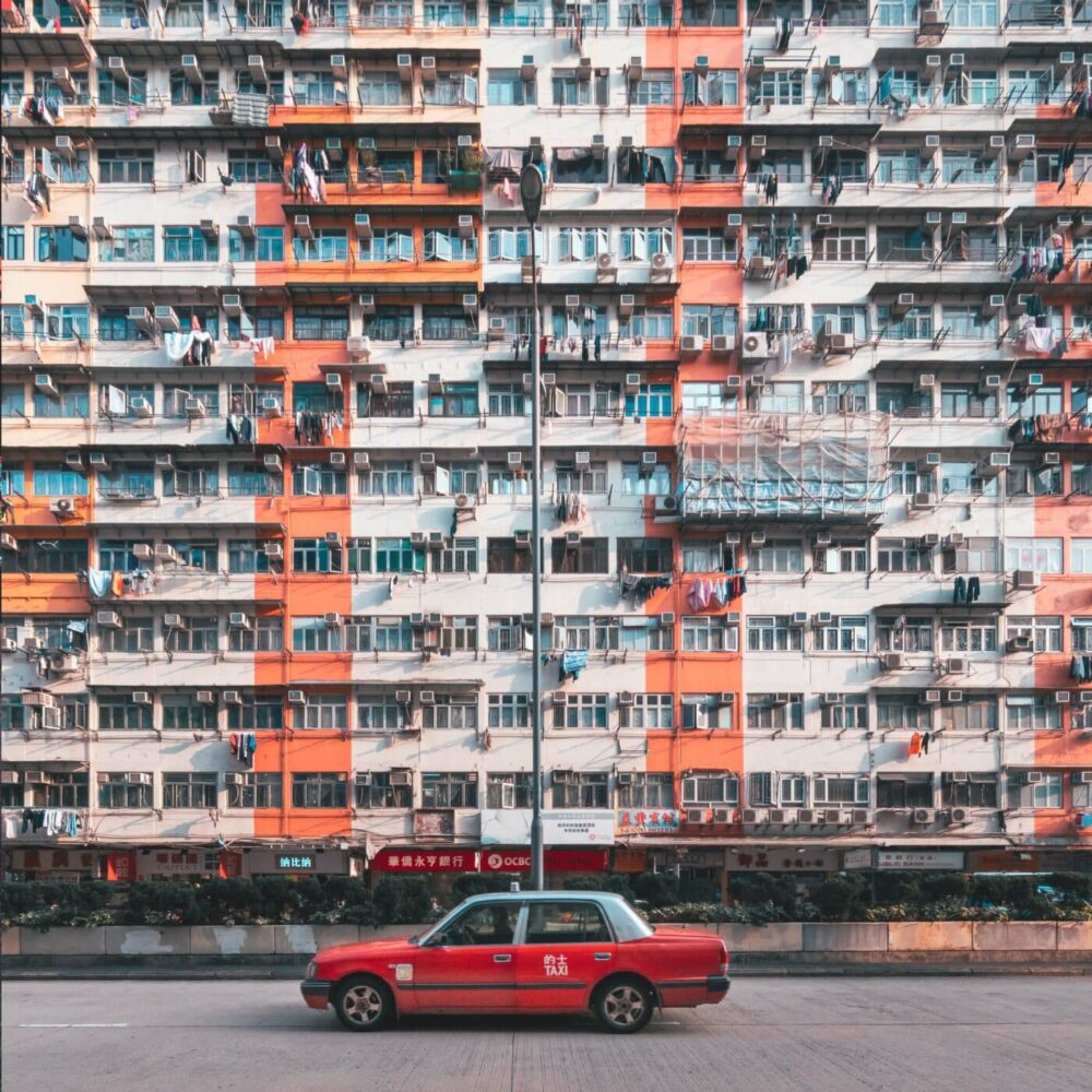 A red taxi is parked by I-Feng Mansion, its vibrant facade a mix of orange, white, and gray. Laundry and air conditioners adorn the many balconies, painting a vivid urban tapestry.