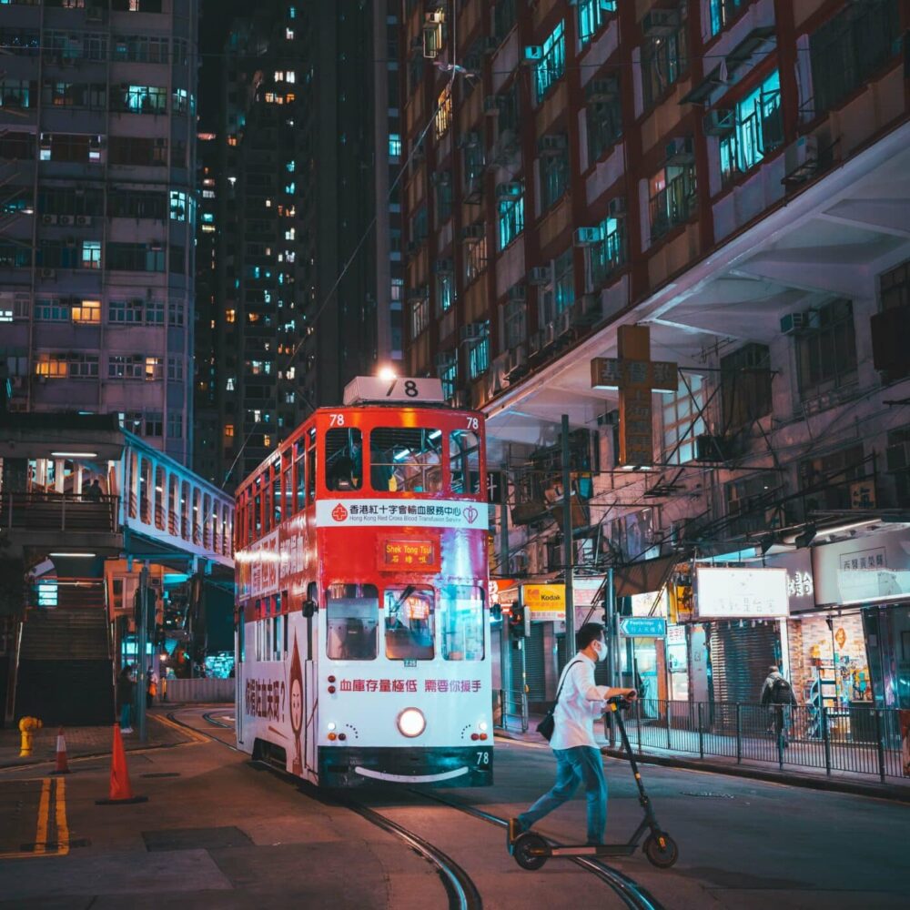 The Red Tram art print captures a nighttime city street scene, featuring a double-decker tram with vibrant lights and signs. A person in a white shirt rides a scooter on the tracks, framed by illuminated urban architecture.